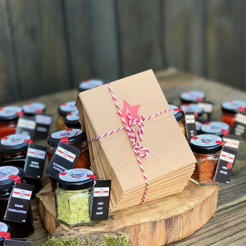 Das Bild zeigt 24 Adventskalender-Tüten mit Schleife drum auf einer Holzscheibe umgeben von verschiedenen Chili Produkten im Glas zum kaufen.