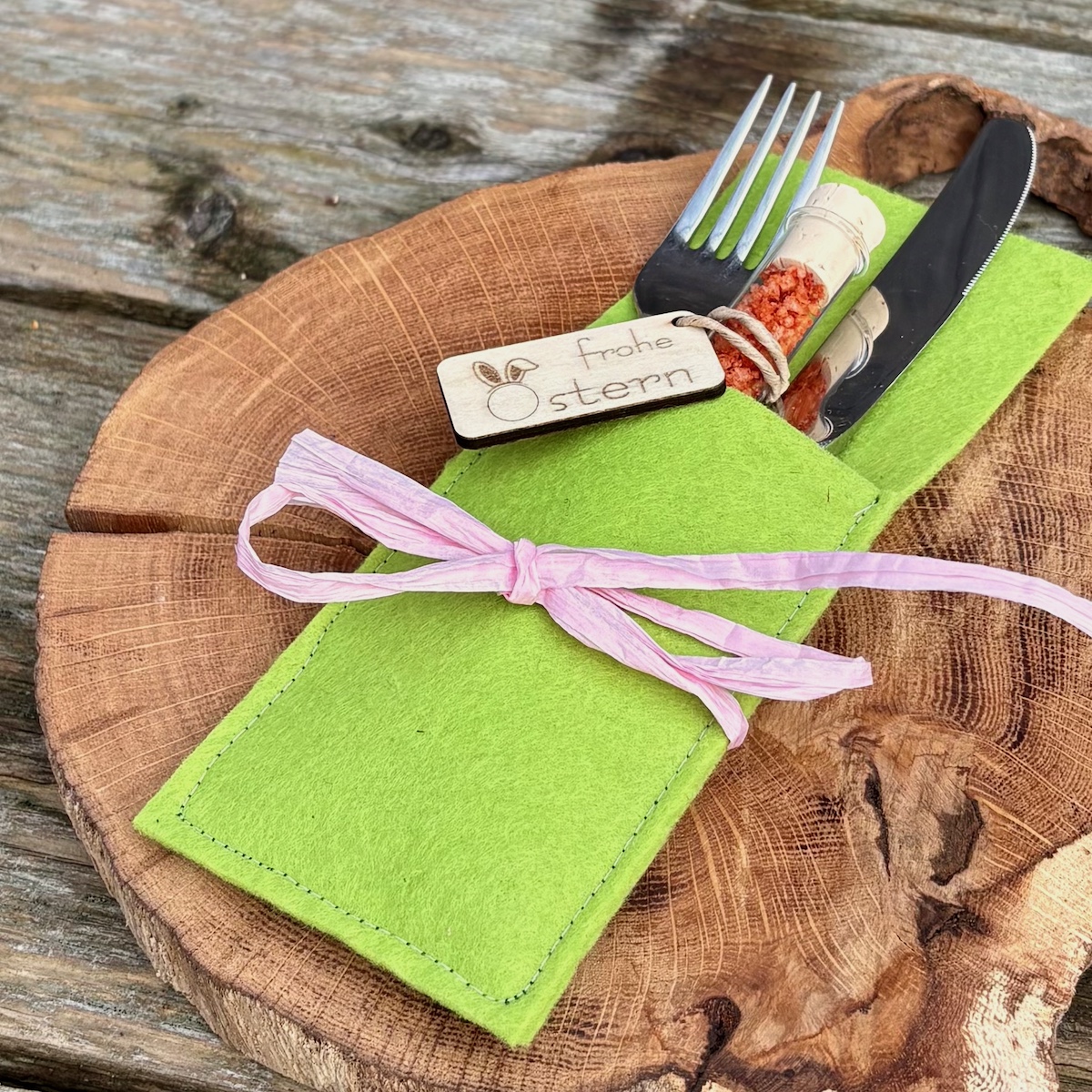 Dekorationsvorschlag Cilisalz smoke im Reagenzglas frohe Ostern in einer Bestecktasche in Frühlingsgrün mit rosa Schleife auf einem Holzteller
