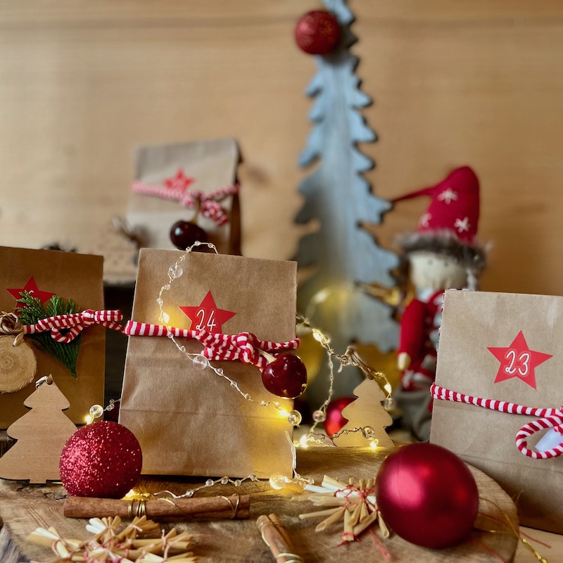 Das Bild zeigt verschiedene Adventskalender-Tütchen auf einer Holzscheibe umgeben von weihnachtlicher Dekoration mit Weihnachtskugeln, Zimtstangen sowie einer Lichterkette. Im Hintergrund sind ein Tannenbaum und eine Weihnachtspuppe zu sehen.