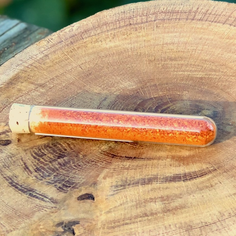 Chili Pulver Heide Staub Kaufen Das Bild zeigt ein Glasröhrchen mit Chili Pulver "Heide Staub"zum Kaufen auf einer Holzscheibe liegend
