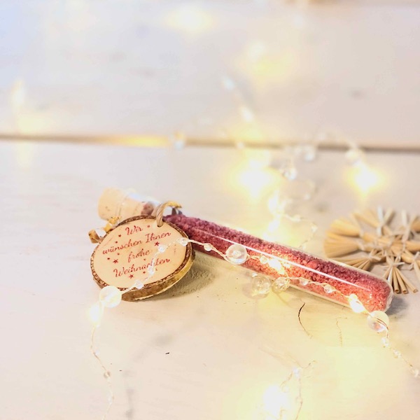 Glasröhrchen mit rotem Blütensalz und Holzanhänger mit der Beschriftung: Wir wünschen Ihnen frohe Weihnachten, als Giveaway .Auf einem Tisch liegend mit Lichterkette und Strohstern