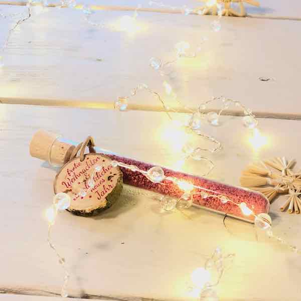 Glasröhrchen mit Blütensalz und Holzanhänger mit dem Aufdruck "frohe Weihnachten und ein glückliches neues Jahr" auf einem Tisch liegend mit Lichterkette und Strohstern