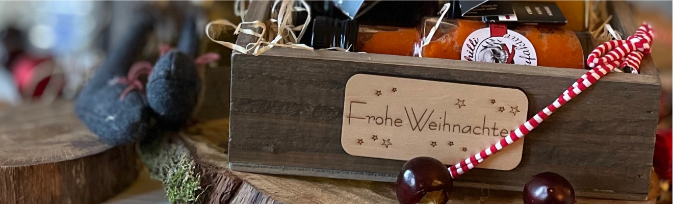 Kulinarische-Geschenke-Weihnachten-als-Giveaway-Kundengeschenk-Kleinigkeit-zu-Weihnachten-kaufen Das Bild zeigt eine Geschenkbox aus Holz mit Schilid Frohe Weihnachten und kulinarische Produkte von der Chilli Manufaktur mit Weihnachtsdekoration