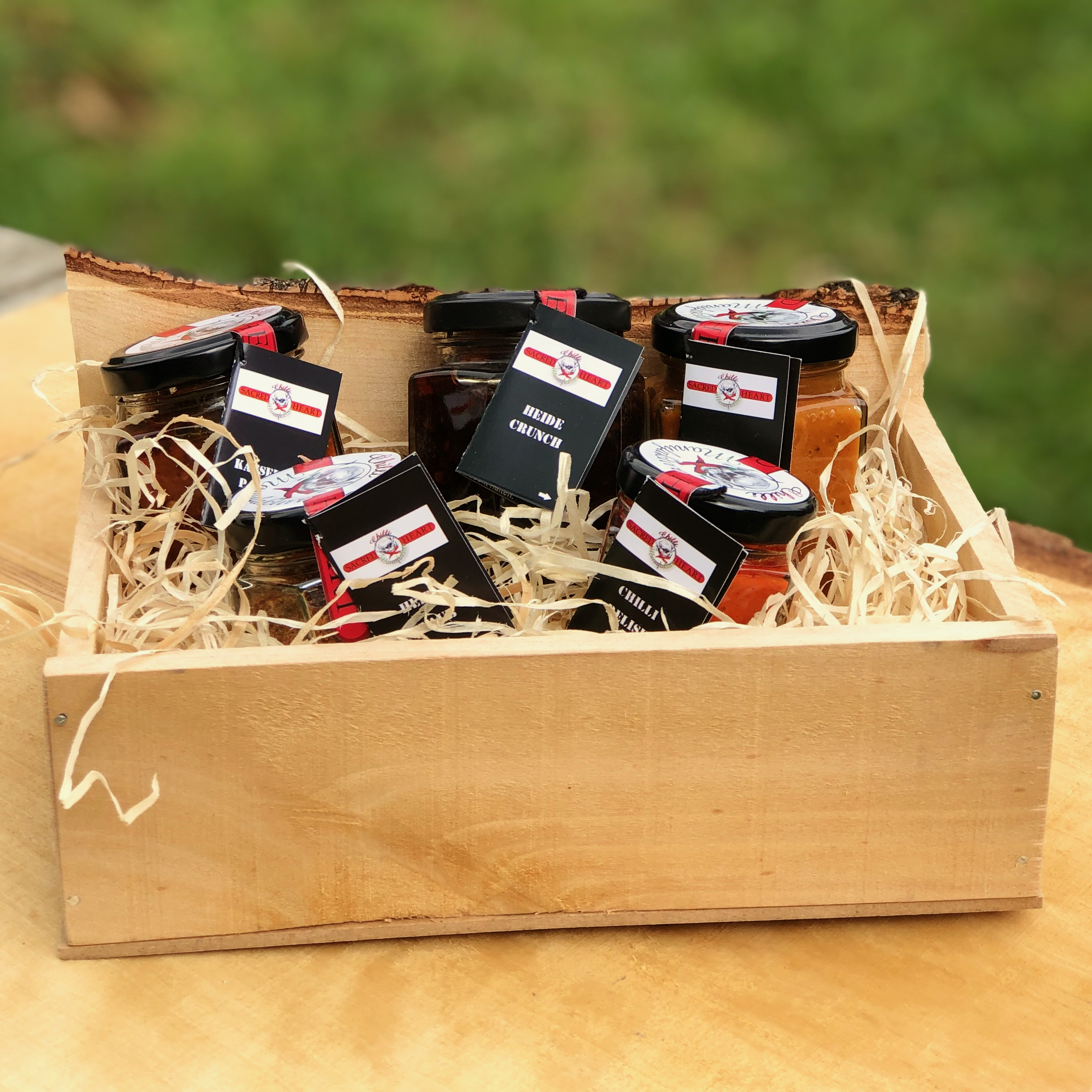 Geschenk Box Heidefeuer Deluxe - Chili Gourmet Box Das Bild zeigt eine Birkenholz-Geschenkbox mit dem Namen Heidefeuer Deluxe -Chili Gourmet Box gefüllt mit Chiliprodukten in Gläsern auf einem Holztisch stehend