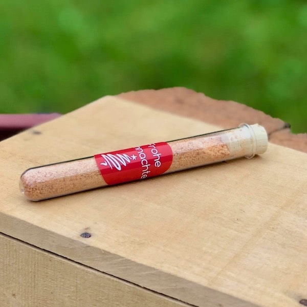 Pommessalz frohe Weihnachten als Tellerdeko zu Weihnachtenkaufen Das Bild zeigt ein Glasröhrchen mit Aufkleber frohe Weihnachten als Tellerdeko zu Weihnachtenauf einem Tisch liegend mit Lichterkette und grünen Zapfen