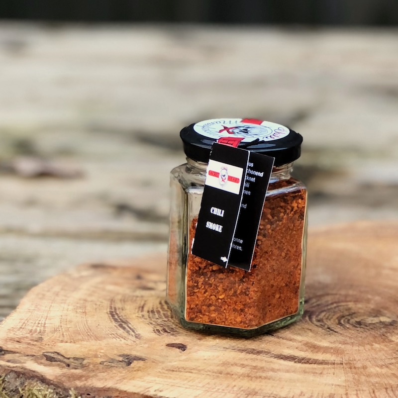 geräuchertes Chili Pulver kaufen geräuchertes Chili Pulver im Glas con der Chilli Manufaktur auf einer Holzscheibe auf einem Tisch stehend