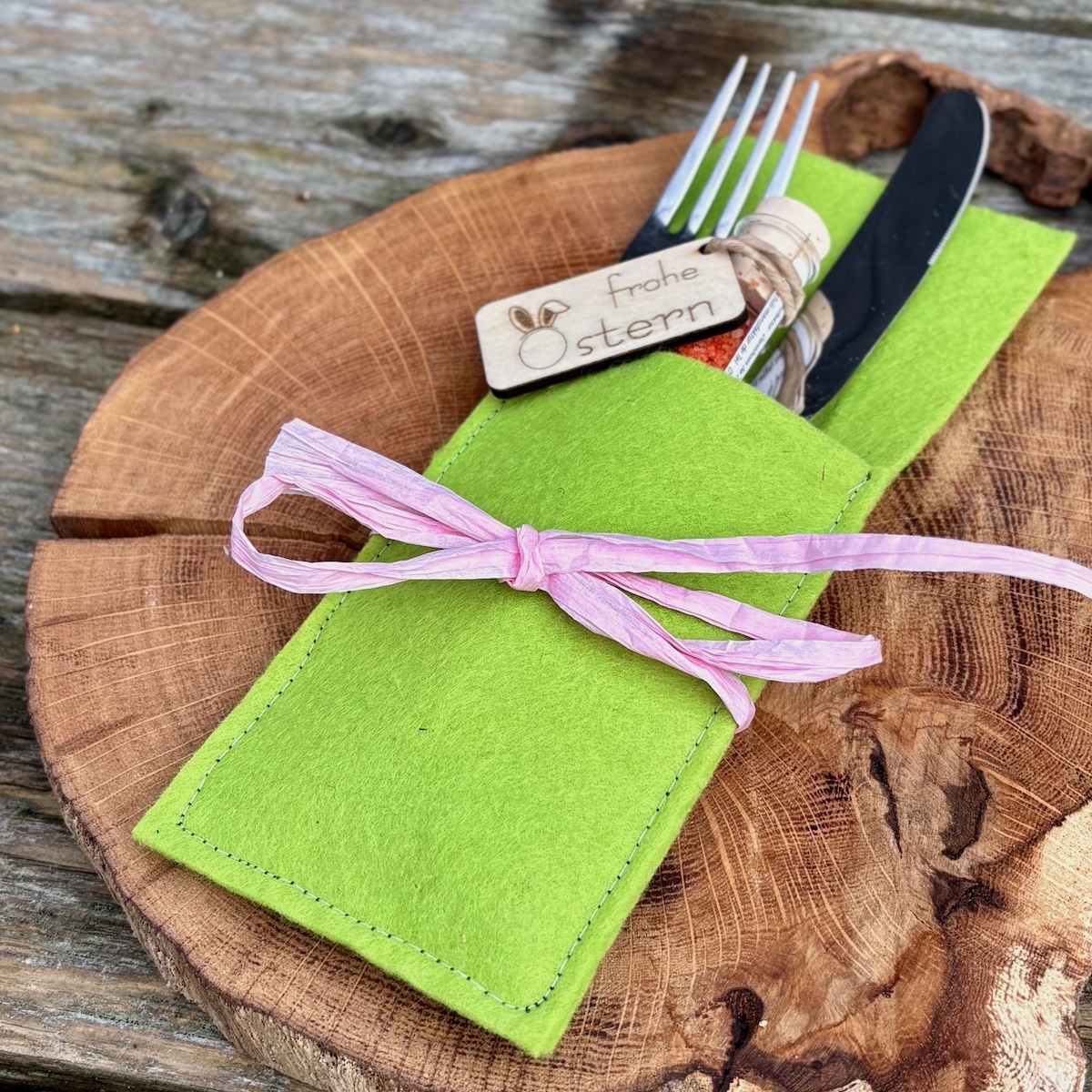 Dekorationsvorschlag Cilisalz frohe Ostern in einer Bestecktasche in Frühlingsgrün mit rosa Schleife auf einem Holzteller