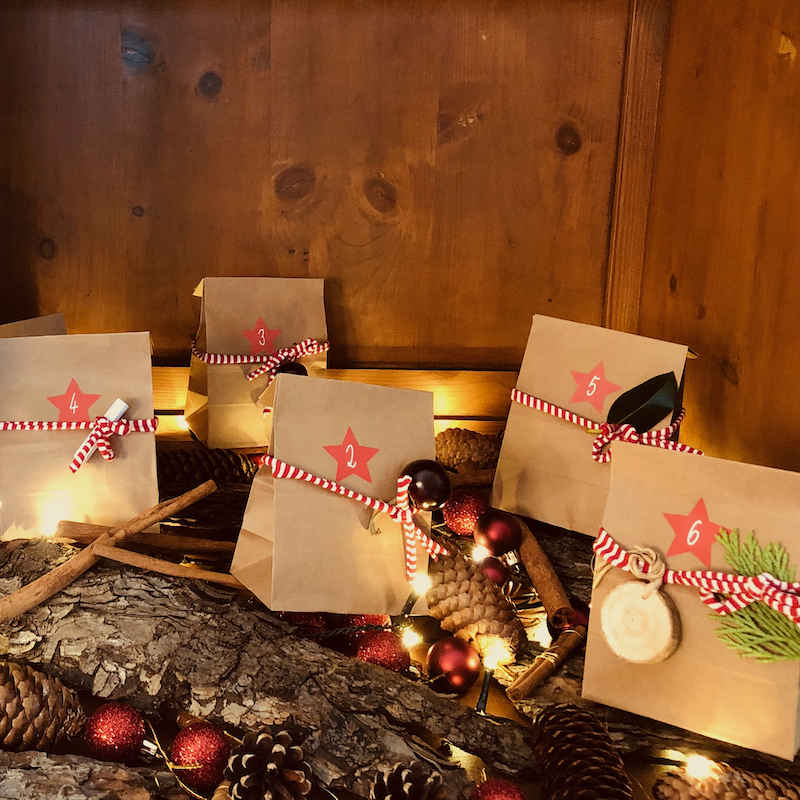 Das Bild zeigt verschiedene Adventskalender-Tütchen präsentiert im inneren eines Schranks auf weihnachtlicher Dekoration mit Weichnachtskugeln, Borke und Tannenzapfen sowie einer Lichterkette.