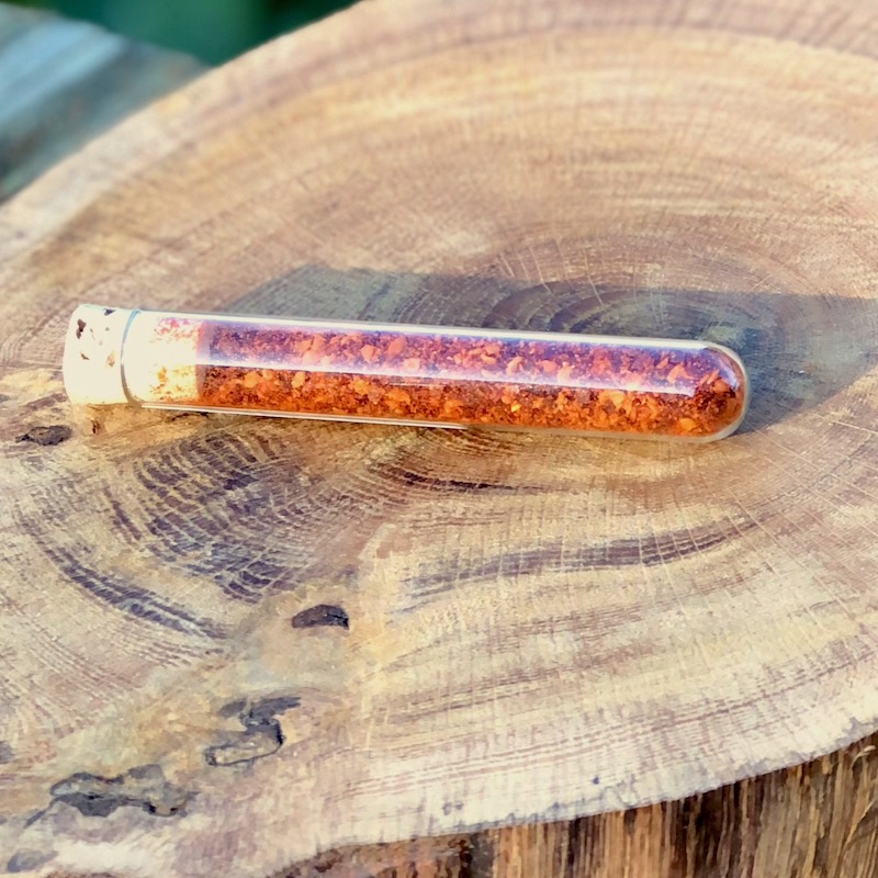 Chili Pulver Schärfegrad 8 kaufen Das Bild zeigt ein Glasröhrchen mit Chili Pulver "Schärfegrad 8" zum Kaufen auf einer Holzscheibe liegend
