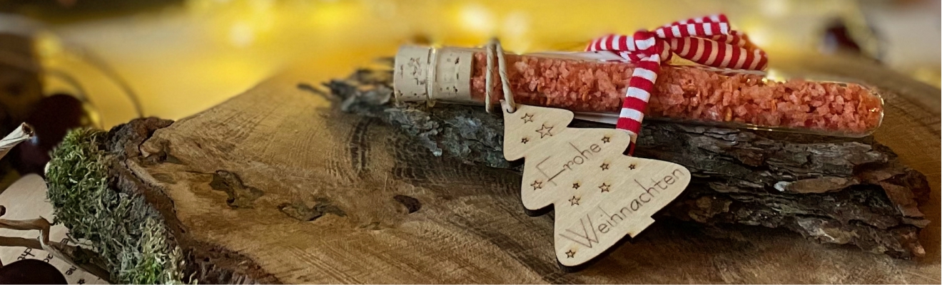 Kleinigkeiten Adventskalender kaufen Das Bild zeigt Chilisalz im Glasröhrchen mit rot weißem Band mit Tannenbaum Anhänger frohe Weihnachten auf einer Holzscheibe liegend mit weihnachtlicher Dekoration als Kleinigkeiten Adventskalender zum kaufen