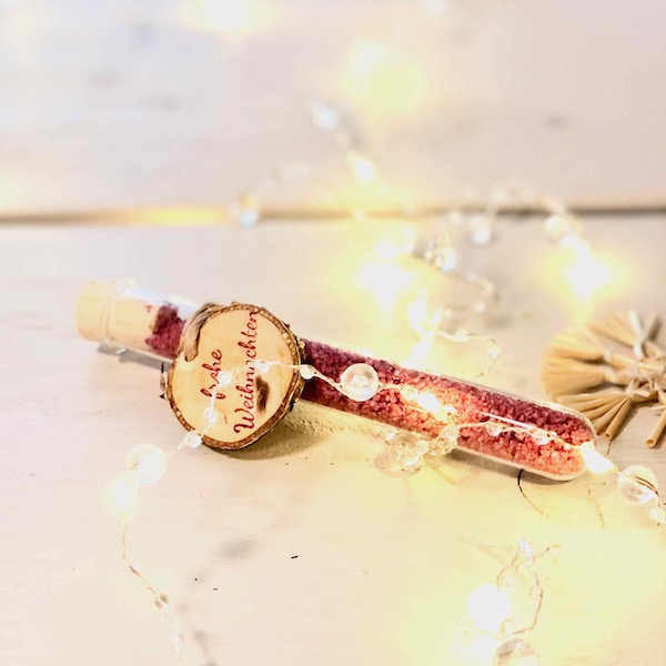 Glasröhrchen mit rotem Blütensalz und Hplzanhänger "frohe Weihnachten" auf einem Tisch liegend mit Lichterkette und Strohstern