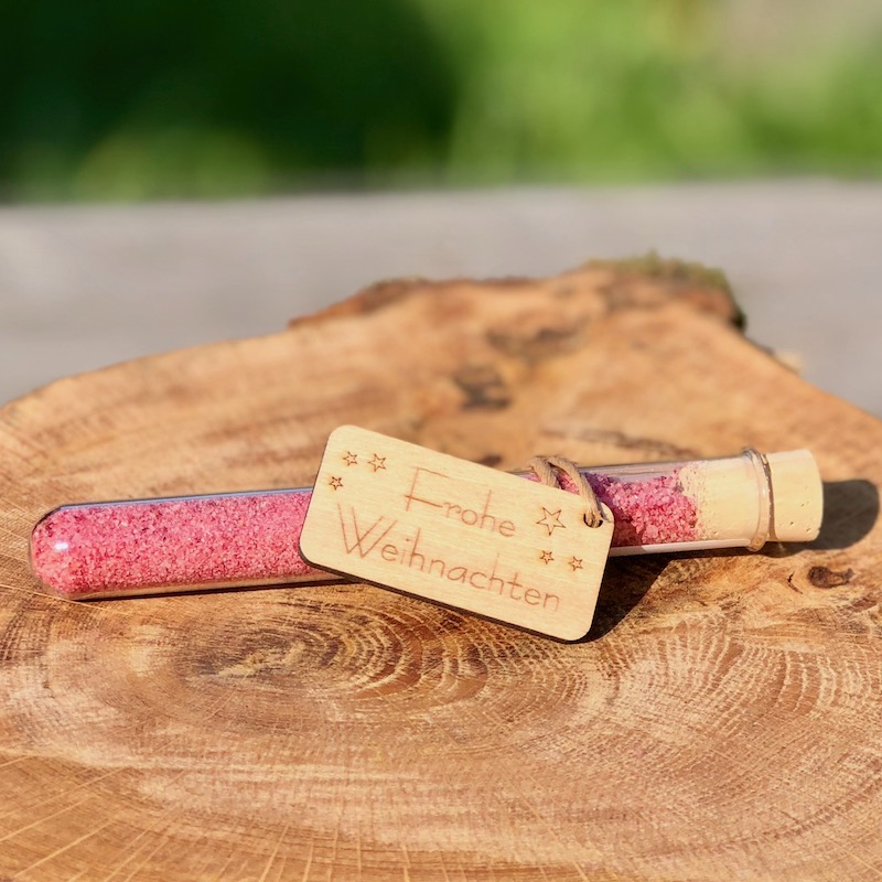 Giveaway frohe Weihnachen Glasröhrchen mit rotem Blütensalz und Hplzanhänger "frohe Weihnachten" auf einem Tisch liegend mit Lichterkette und Strohstern