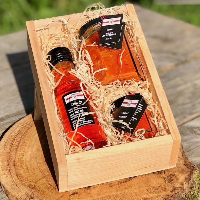 Das Bild zeigt die Chili Geschenkbox feuriger Start. Es ist eine Holzbox mit dem inhalt Chili Brotaufstrich mild, chiliöl und Chili Relish rot