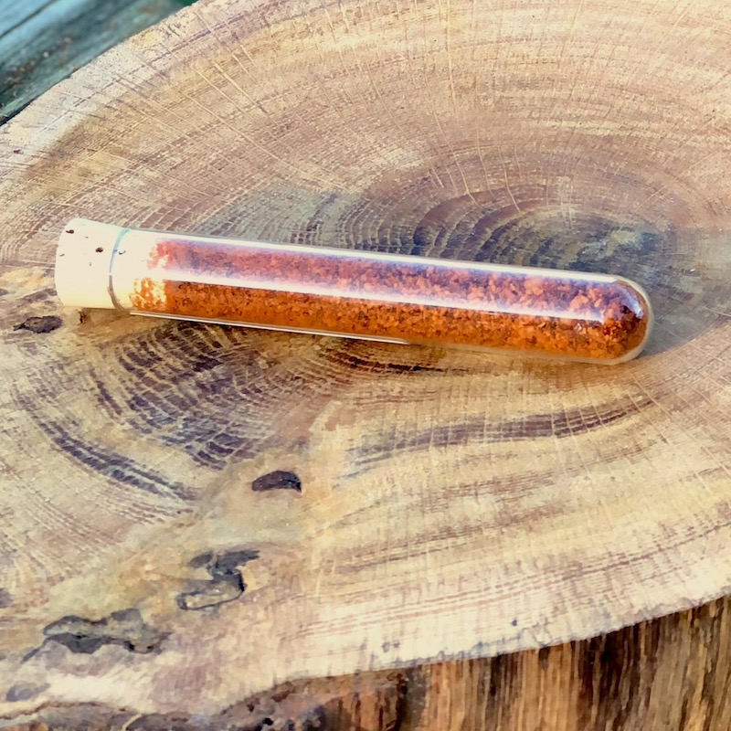 Geräuchertes Chili Pulver kaufen Das Bild zeigt ein Glasröhrchen mit geräucherten Chili Pulver "Schärfegrad 6" zum Kaufen auf einer Holzscheibe liegend